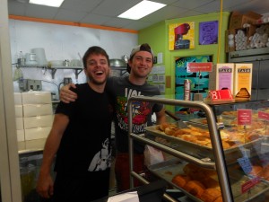 Two men standing behind the counter at a shop with various pastries in the clear glass cover of the counter