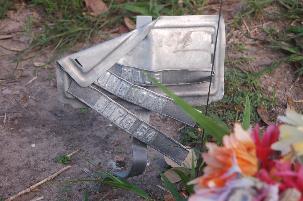 A bent metal burial marker with the words ""Male, Unknown, 417654" falling off