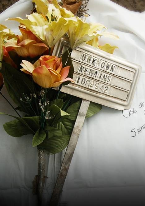 Metal grave marker with "Unknown Remains" on it with orange and yellow flowers over a white body bag
