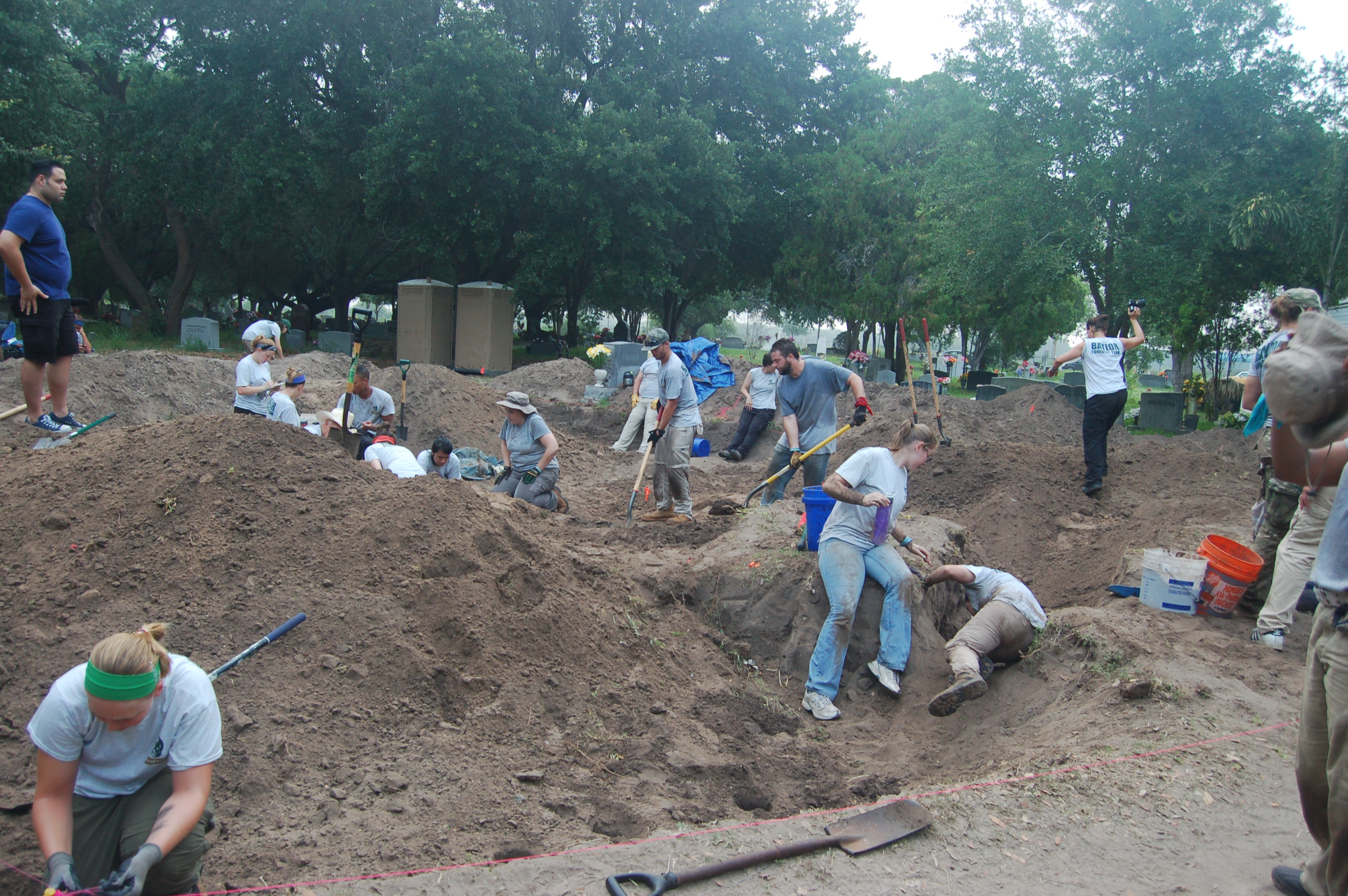 Overview pictures of multiple burials being worked on by many individuals with mounds of dirt surrounding