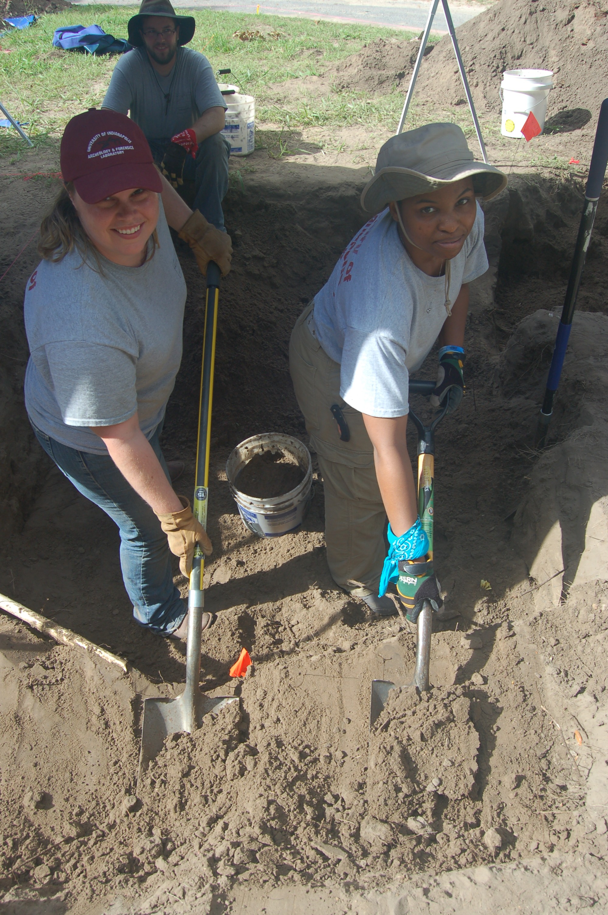 UIndy team members smiling at the camera while shoveling inside a burial