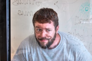 Team member Justin standing behind a clear dry-erase board with a mustache and monocle drawn on it