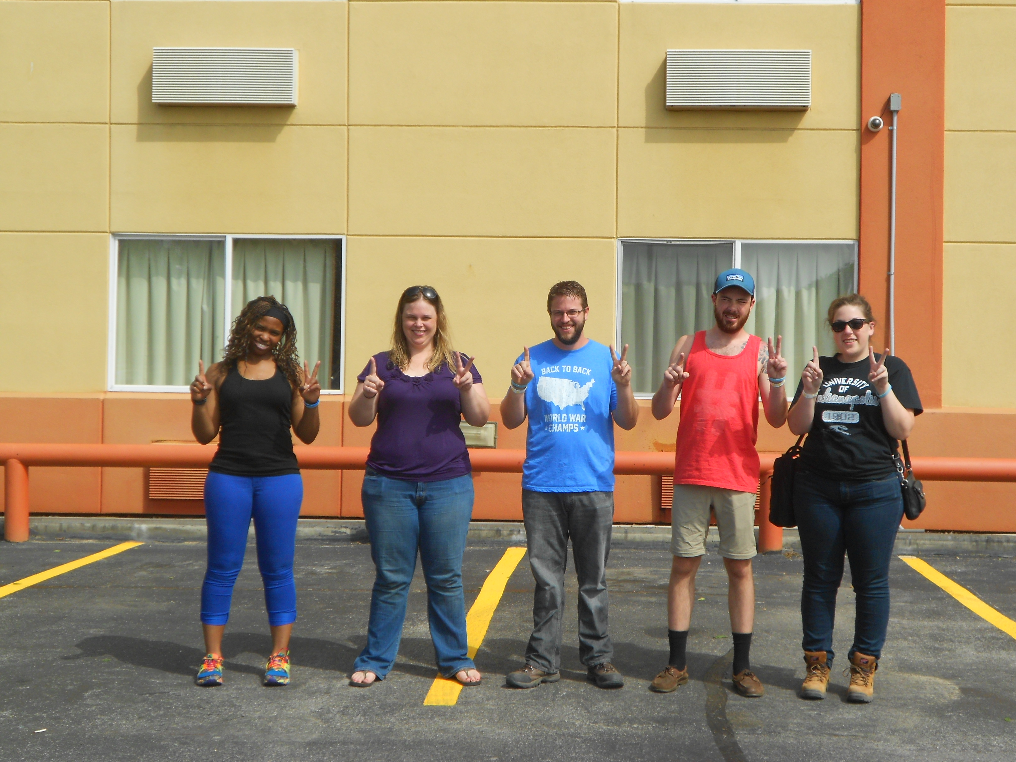 Group picture outside a yellow hotel with one finger raised on one hand and two fingers raised on the other hand