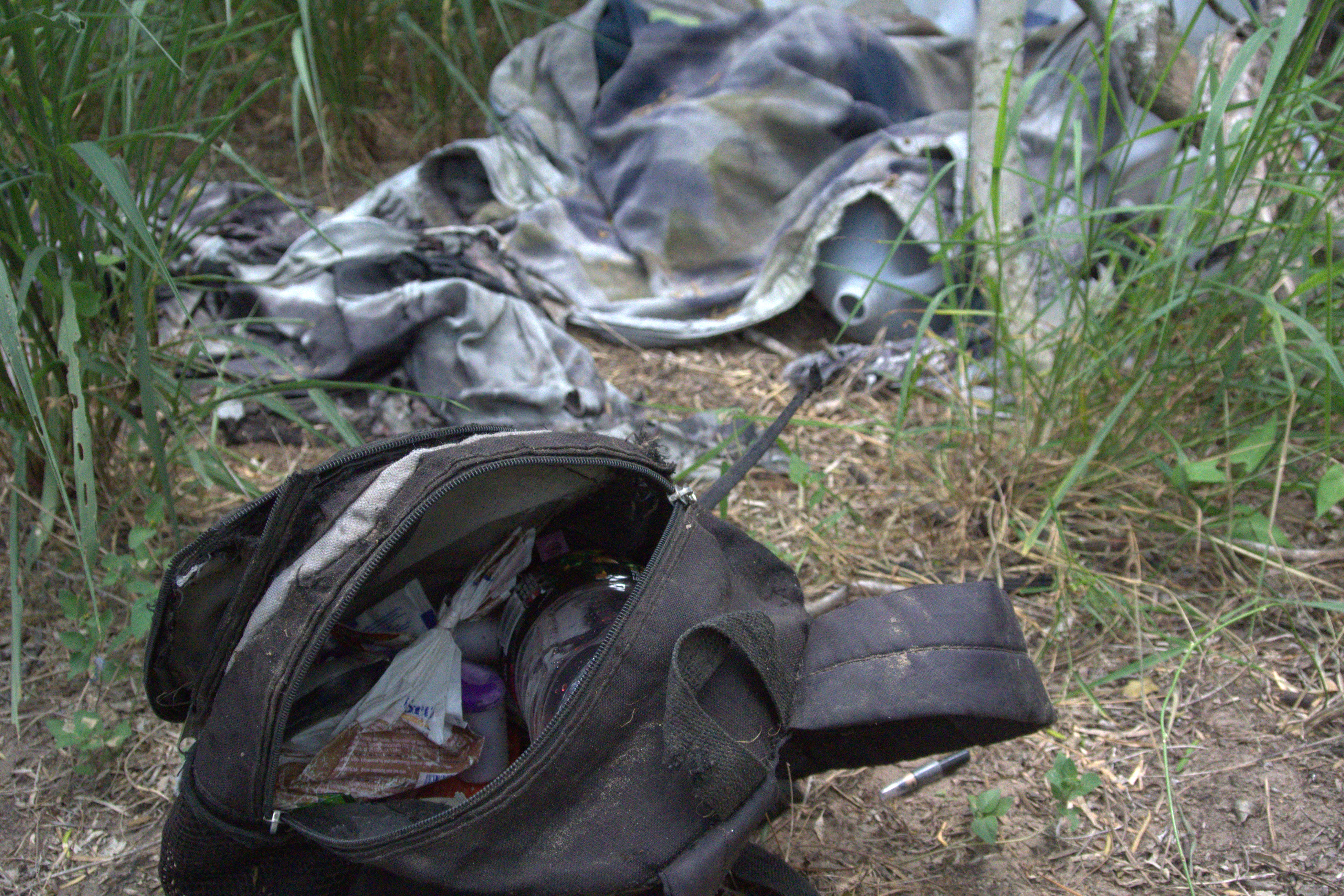 A discarded backpack, torn clothes, and a water jug in the brush