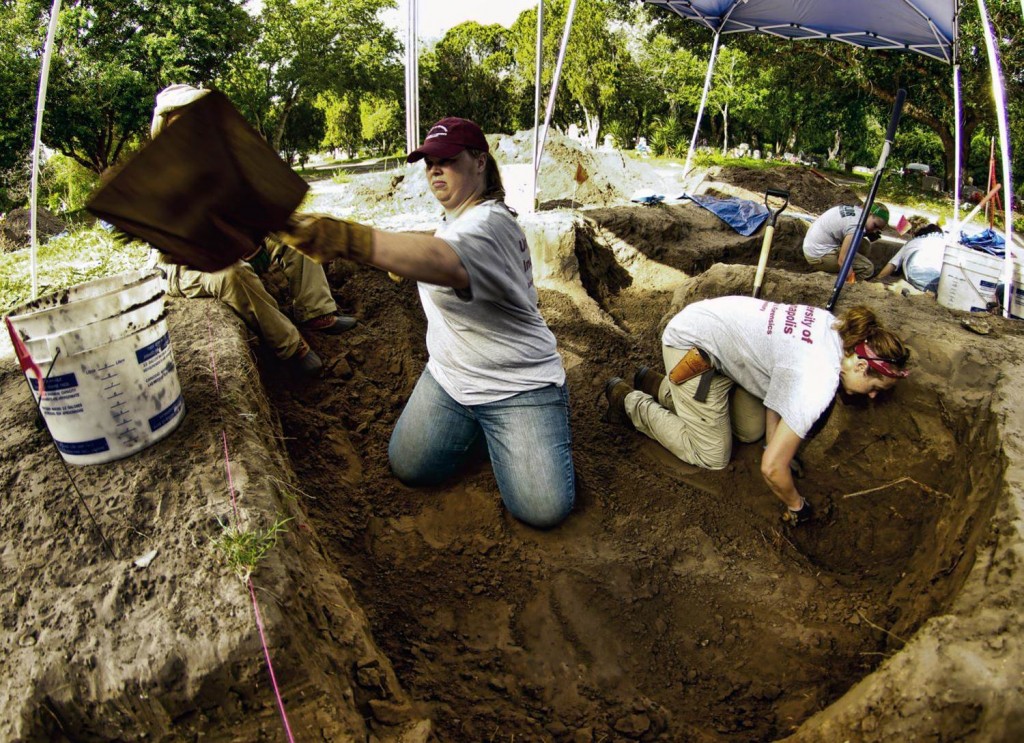 team members working in multiple burials under a tent, scooping out dirt with shovels into buckets on the ground level. Others using trowels and more tools around on the ground level