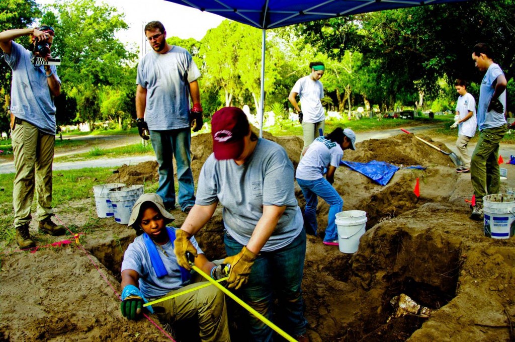 Ground surface picture over members crossing tape measures to map in a burial, while others take pictures or continue working in the background