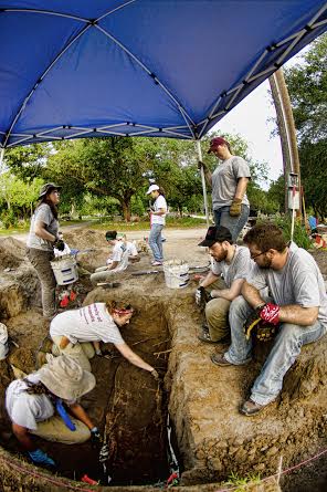 Team members working within a burial under a shade tent while others sit above