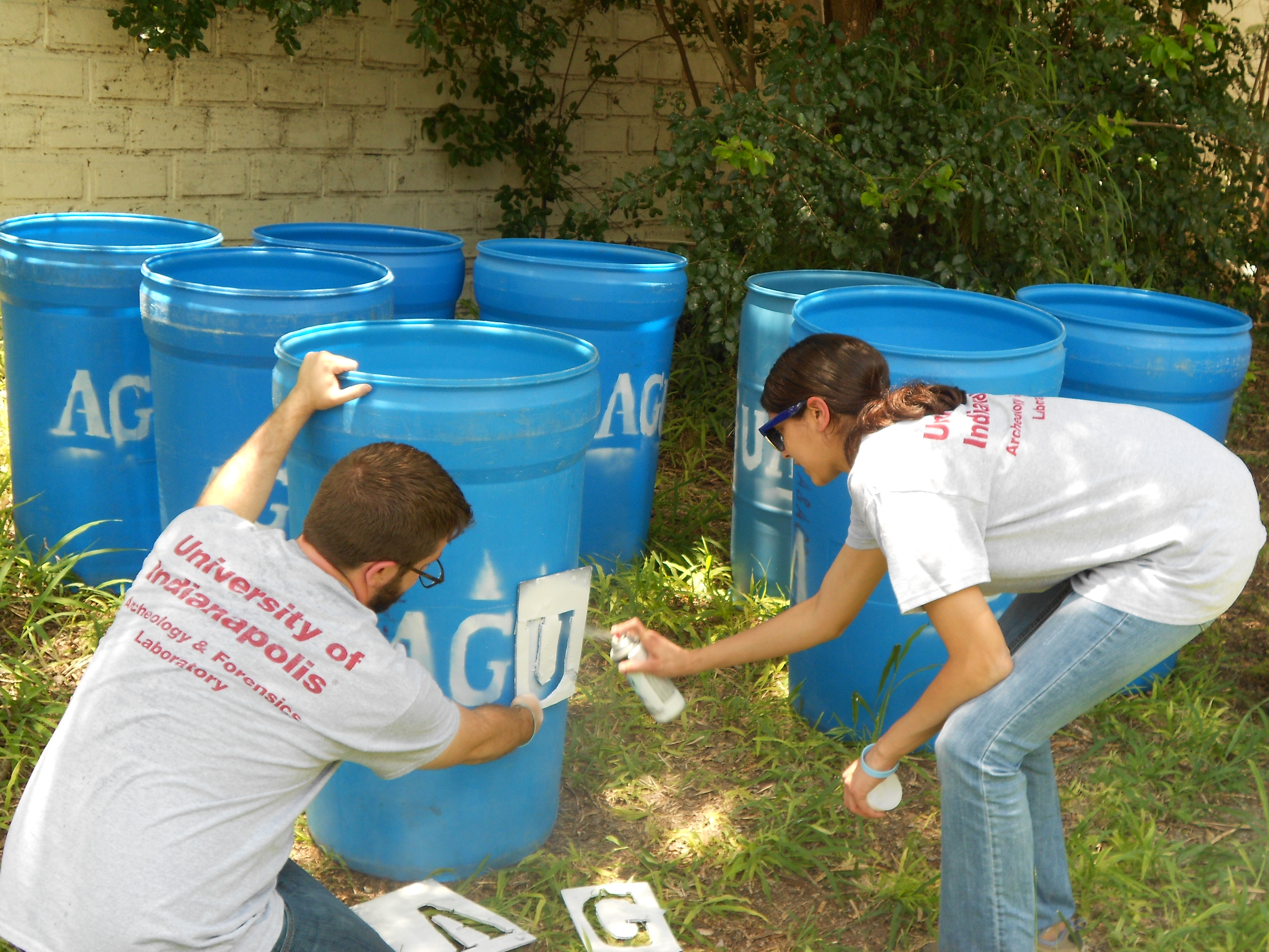 Team members spray painting a "U" with a stencil on a water station barrel outside with multiple barrels in the background