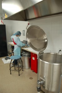 Student in gown, hair net, and gloves next to the large processing kettle in the processing suite