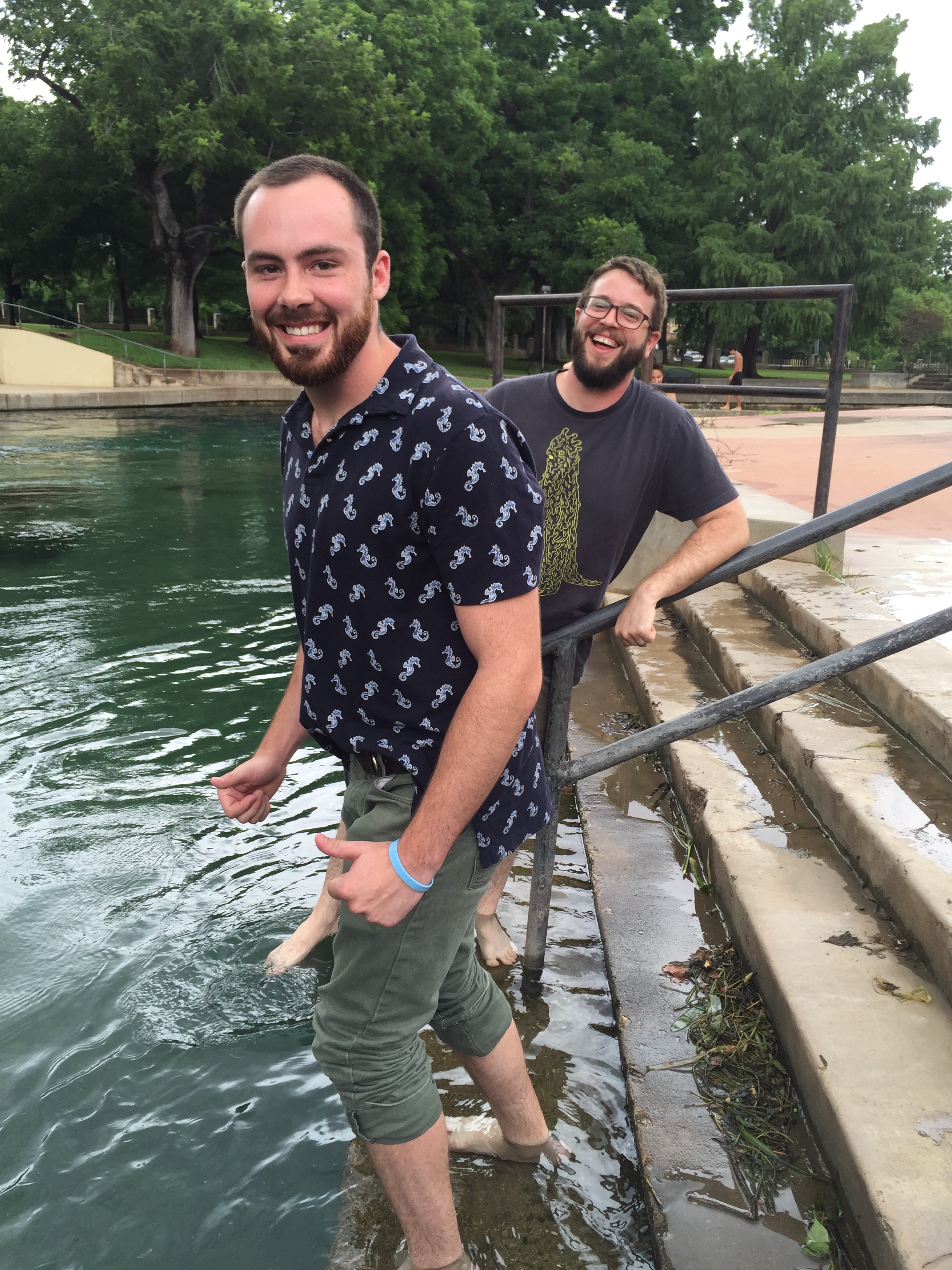 Two Beyond Borders team members giving smiles to the camera as they dip their feet in the San Marcos River on a stair platform leading from the sidewalk into the river