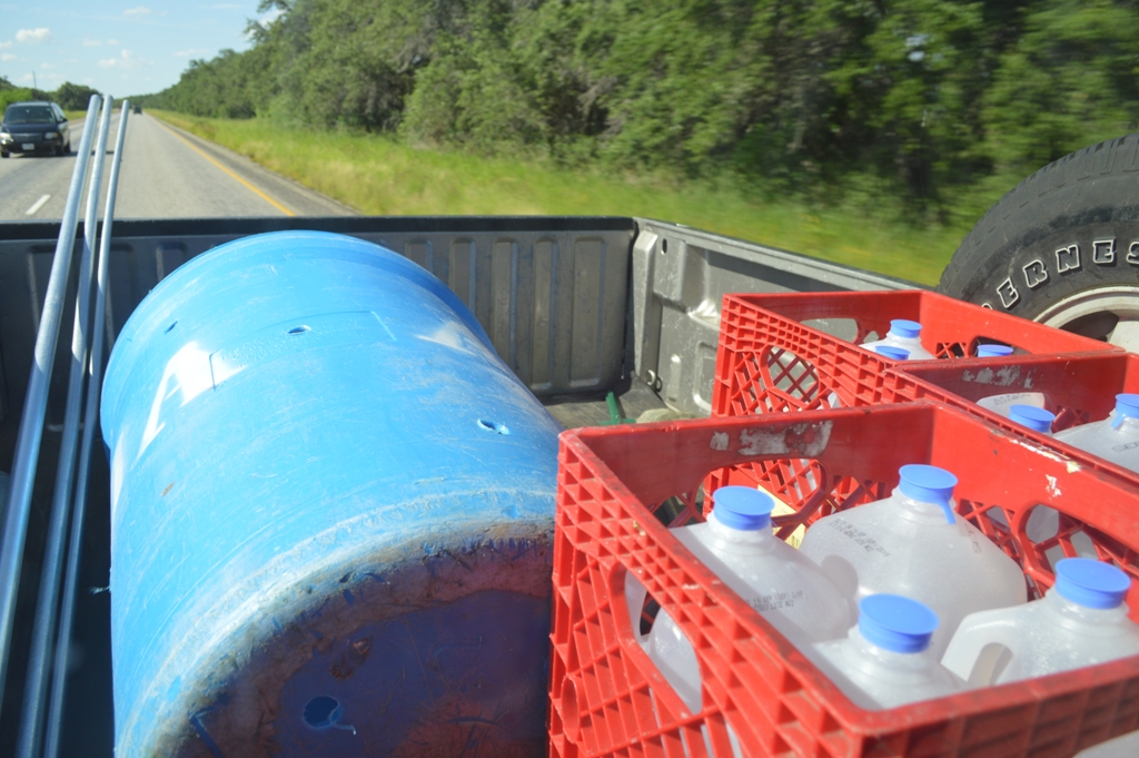 A large blue barrel and milk carts of gallon water jugs in the bed of a truck.