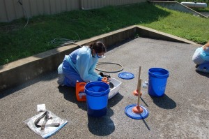 A person in PPE kneeling on the ground with buckets, plungers, and a plastic tub