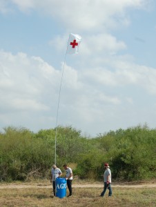 Three people around a blue barrel with a red cross flag flying in the sky