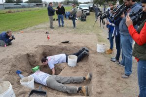 Media standing over and filming two team members excavating a burial.