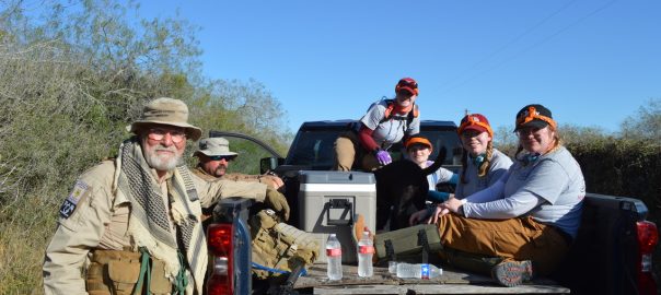 Back of a truck with 4 individuals and a dog sitting in the bed of the truck, two men stand next to the truck