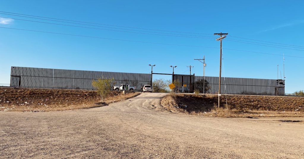 View of the US-Mexico border wall, with an open gate and two Border Patrol vehicles.